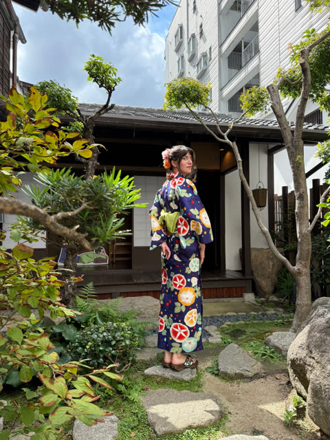 A woman in a blue floral kimono stands on a stone path in a Japanese garden and looks over her shoulder.