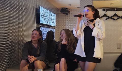 A woman sings into a microphone while two other women sit and watch her inside a karaoke room.