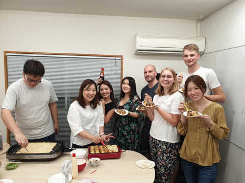A group of eight people smile while gathered around a table cooking and eating takoyaki together in an indoor room.