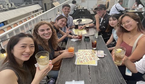 A group of eight people holding drinks smile at the camera while sitting at a wooden table on an outdoor rooftop patio.