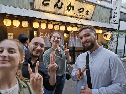 Four people smile and make peace signs while taking a selfie in front of a Japanese restaurant with paper lanterns.
