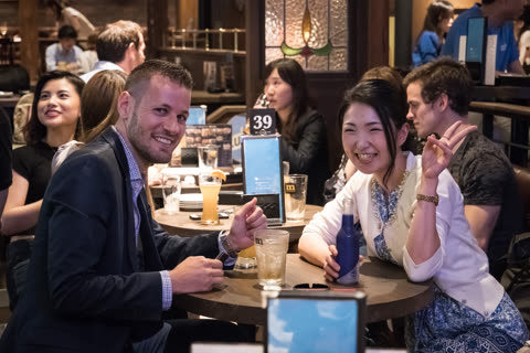 A man and a woman smile at a table in a busy pub, where the woman holds up a peace sign.