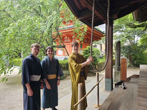 Three men in traditional robes stand at a Japanese temple, with one man holding a large rope of hanging wooden beads.