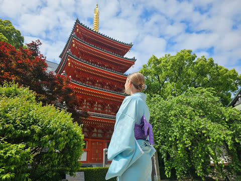 A person in a light blue kimono stands looking up at a multi-storied red pagoda surrounded by trees.